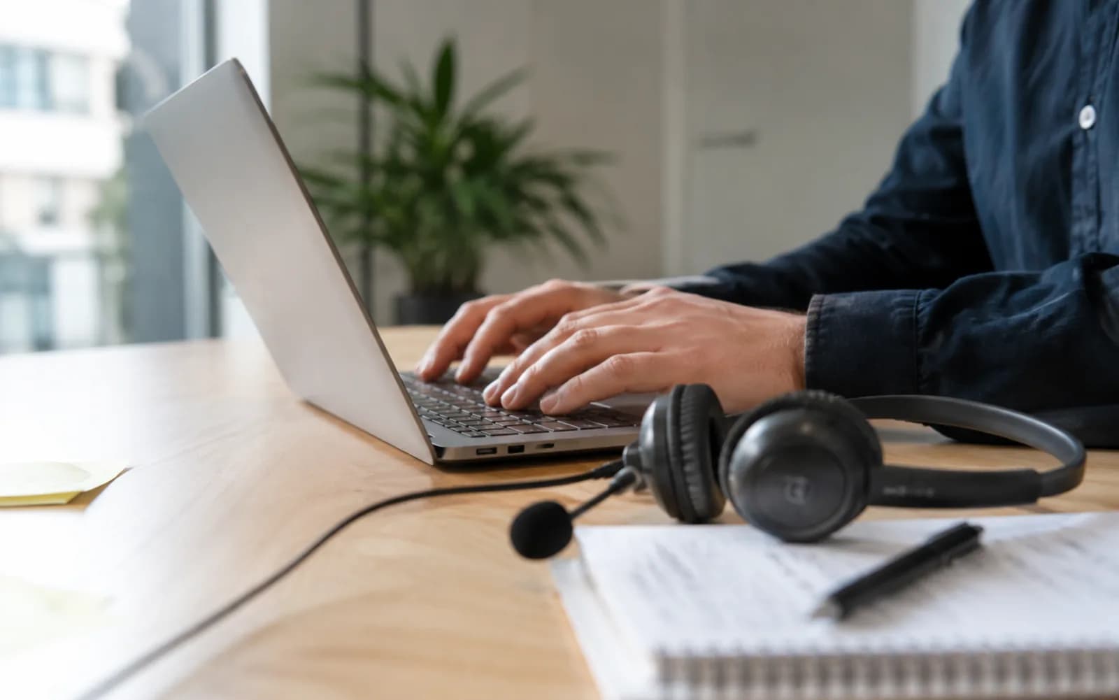 Hände an einem Laptop auf hellem Holz-Schreibtisch, Headset und Notizblock daneben — ruhige Büro-Atmosphäre